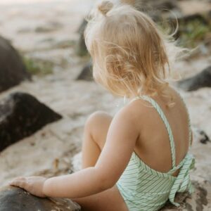 A little girl in the Retro Wave By Ina - Mara One-Piece - Fern Stripe sitting on rocks at the beach.