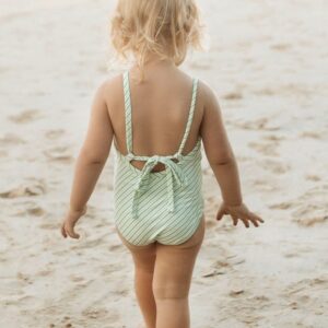 A little girl in a Retro Wave By Ina - Mara One-Piece - Fern Stripe swimsuit walking on the beach.