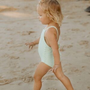 A little girl in a Retro Wave By Ina - Mara One-Piece - Fern Stripe swimsuit walking on the beach.