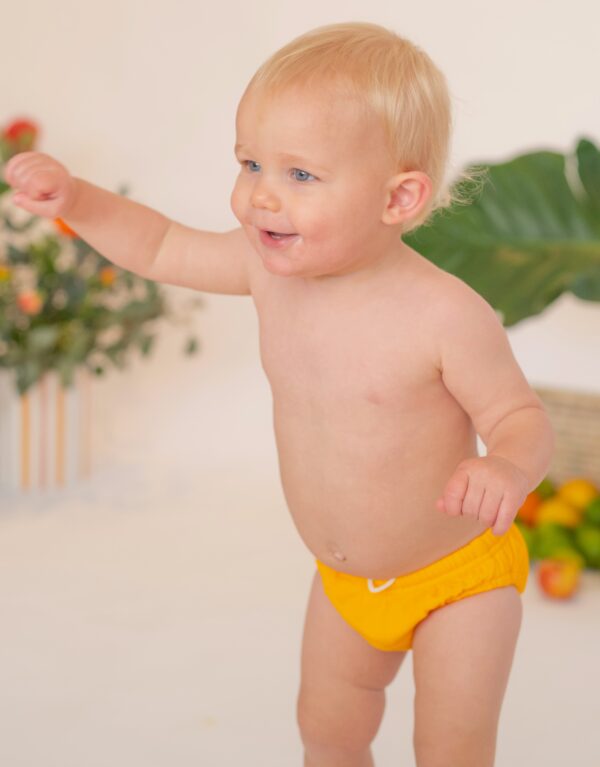 A baby in a Retro Wave By Ina - Lumi Brief Swim Nappy standing in front of a plant.