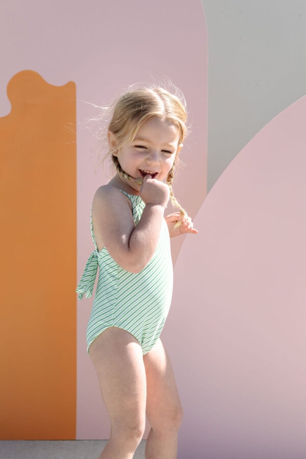 Young child in a Mara One-Piece - Fern Stripe swimsuit standing in front of a colorful backdrop, looking pensive.