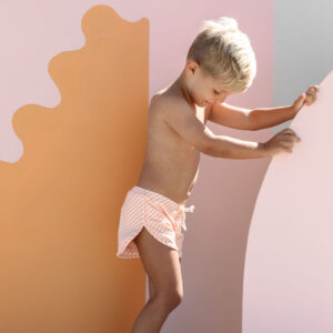 A young child in Mesa Trunks - Marigold Stripe stands between colorful cut-out panels, looking down and touching the wall with one hand.