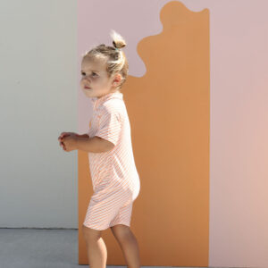 A young child wearing a Zimmi Onesie - Marigold Stripe stands in profile against a pastel-colored background with abstract shapes.