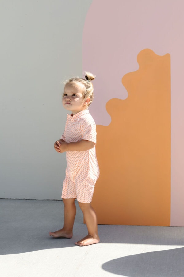 A young child in a Zimmi Onesie - Marigold Stripe standing in front of a colorful abstract backdrop.