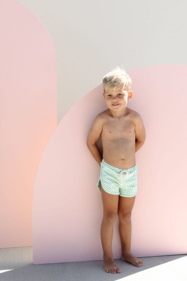 Young boy in Mesa Trunks - Fern Stripe standing in front of a pink curved wall.