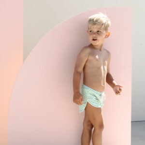 A young child in Mesa Trunks - Fern Stripe swim shorts standing beside a pink curved panel in a brightly lit setting.