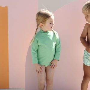 Two young children standing against a colorful backdrop, one dressed in a Nella Rash Shirt - Fern and the other in swim shorts.