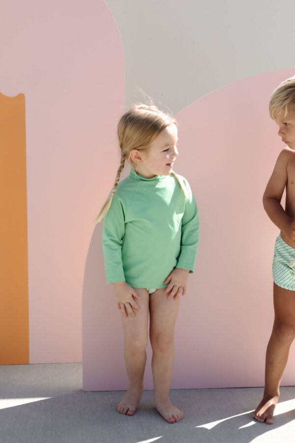 Two young children standing against a colorful backdrop, one dressed in a Nella Rash Shirt - Fern and the other in swim shorts.