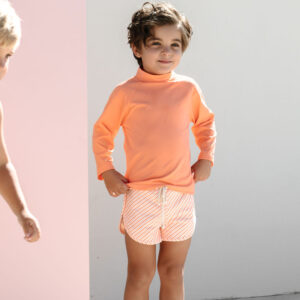 Child in Nella Rash Shirt - Marigold and striped shorts standing confidently with hands on hips.