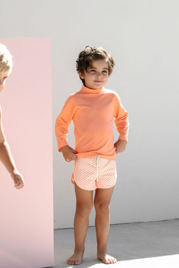 Child in Nella Rash Shirt - Marigold and striped shorts standing confidently with hands on hips.