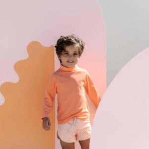A young child wearing a Nella Rash Shirt - Marigold and patterned shorts stands smiling against a pastel backdrop with curvy shapes.