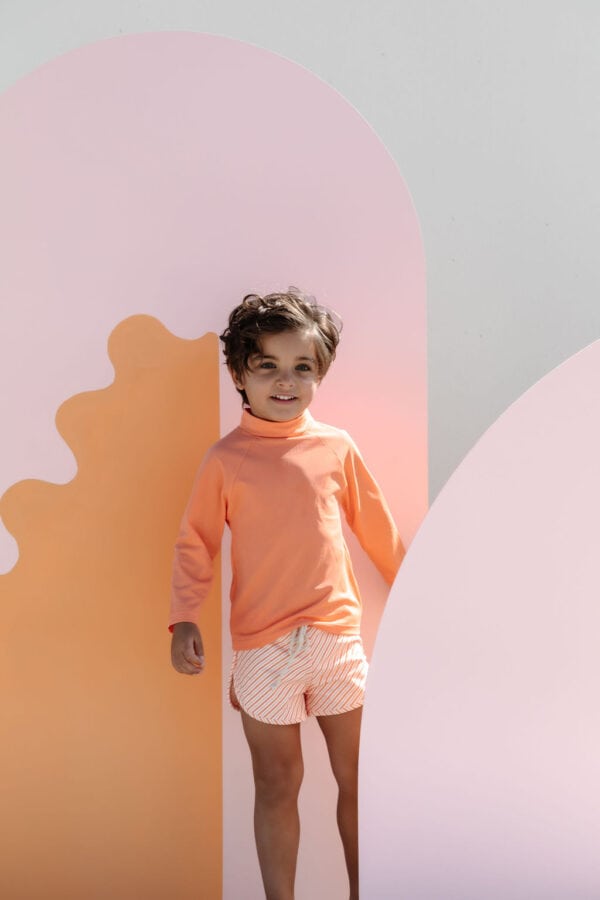 Child in a peach-colored sweatshirt and Mesa Trunks - Marigold Stripe standing in front of a pink and cream abstract backdrop.