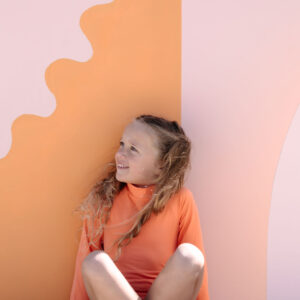 A young girl in a June One-Piece - Marigold sitting and smiling against a backdrop with playful pink and orange shapes.