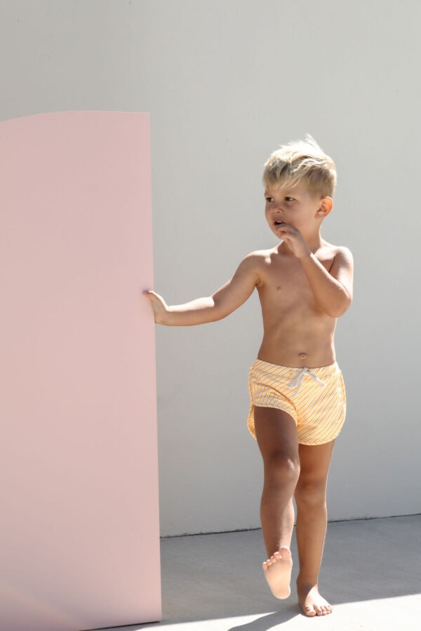 A curious child in Mesa Trunks - Dandelion Stripe swimwear standing beside a pink panel with a thoughtful expression.