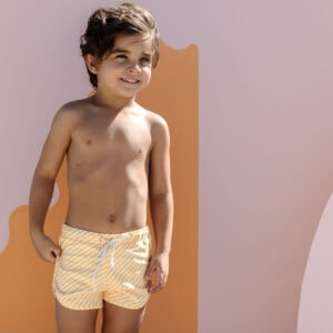 A young child smiling and wearing Mesa Trunks - Dandelion Stripe against a pastel background.