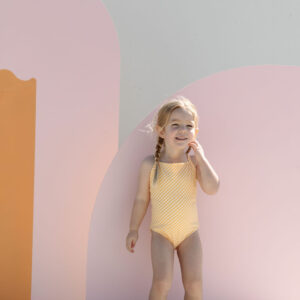 Young child in a Mara One-Piece - Dandelion Stripe swimsuit standing in front of a colorful backdrop with pink and yellow arches.