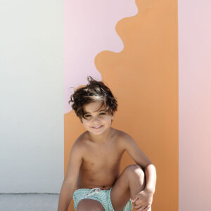 A young child in Mesa Trunks - Fern Stripe swim shorts smiling while kneeling in front of a colorful backdrop.