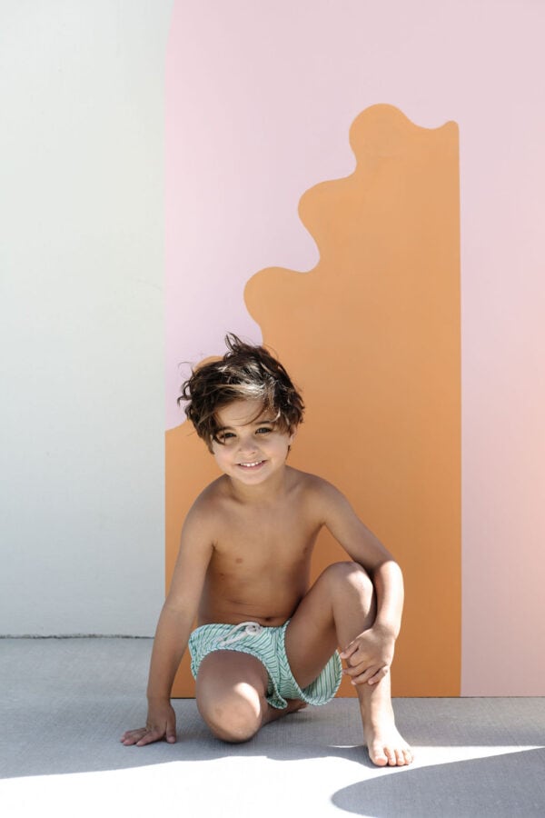 A young child in Mesa Trunks - Fern Stripe swim shorts smiling while kneeling in front of a colorful backdrop.