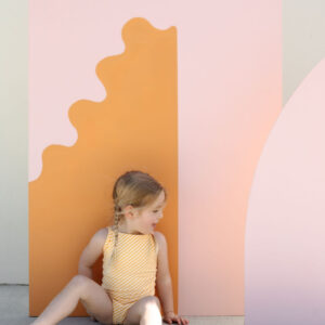 A young child sits beside Mara One-Piece - Dandelion Stripe on a bright background.