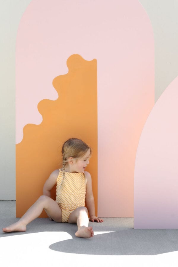 A young child sits beside Mara One-Piece - Dandelion Stripe on a bright background.
