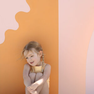 A young girl with braided hair sitting and smiling against a playful backdrop with wavy pastel pink and orange panels in the Mara One-Piece - Dandelion Stripe.