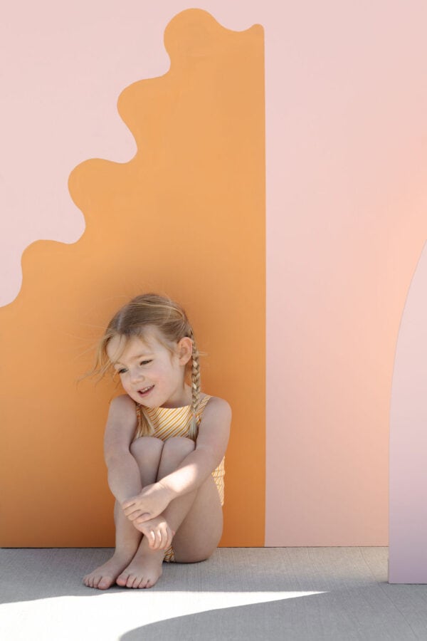 A young girl with braided hair sitting and smiling against a playful backdrop with wavy pastel pink and orange panels in the Mara One-Piece - Dandelion Stripe.