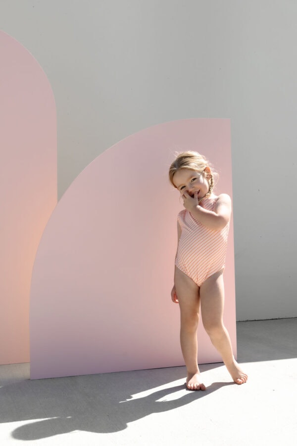 A child in a Mara One-Piece - Marigold Stripe swimsuit standing beside a pink curved backdrop with a playful expression.