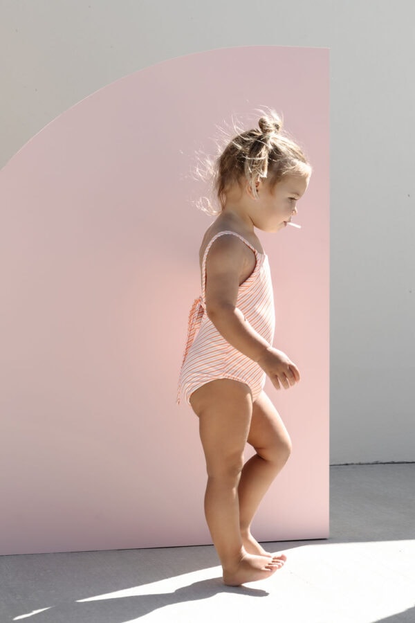 A toddler in a Mara One-Piece - Marigold Stripe swimsuit stands beside a pink curved backdrop.