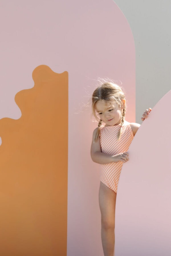 A young girl in a Mara One-Piece - Marigold Stripe swimsuit peeking around a colorful abstract backdrop.