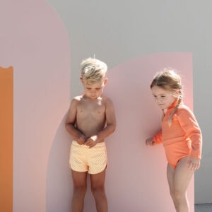 Two children in swimwear standing beside a colorful backdrop, the boy looking down and the girl leaning in with a curious expression, wearing the June One-Piece - Marigold.