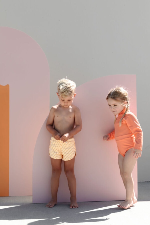 Two children in swimwear standing beside a colorful backdrop, the boy looking down and the girl leaning in with a curious expression, wearing the June One-Piece - Marigold.