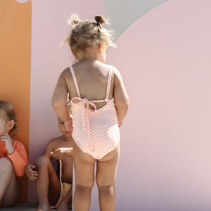 A toddler in a Mara One-Piece - Marigold Stripe standing in front of a wall with abstract design, while other children sit beside.