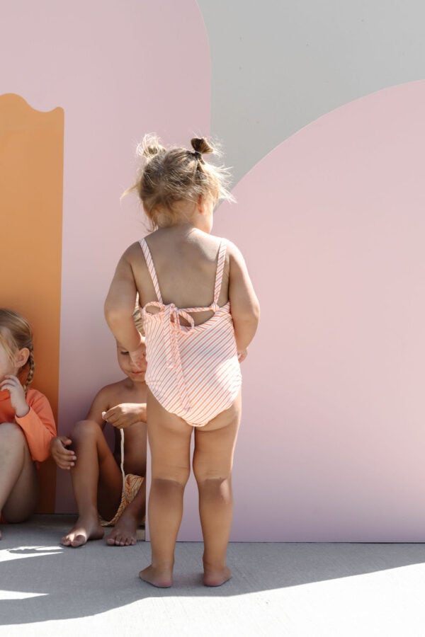 A toddler in a Mara One-Piece - Marigold Stripe standing in front of a wall with abstract design, while other children sit beside.