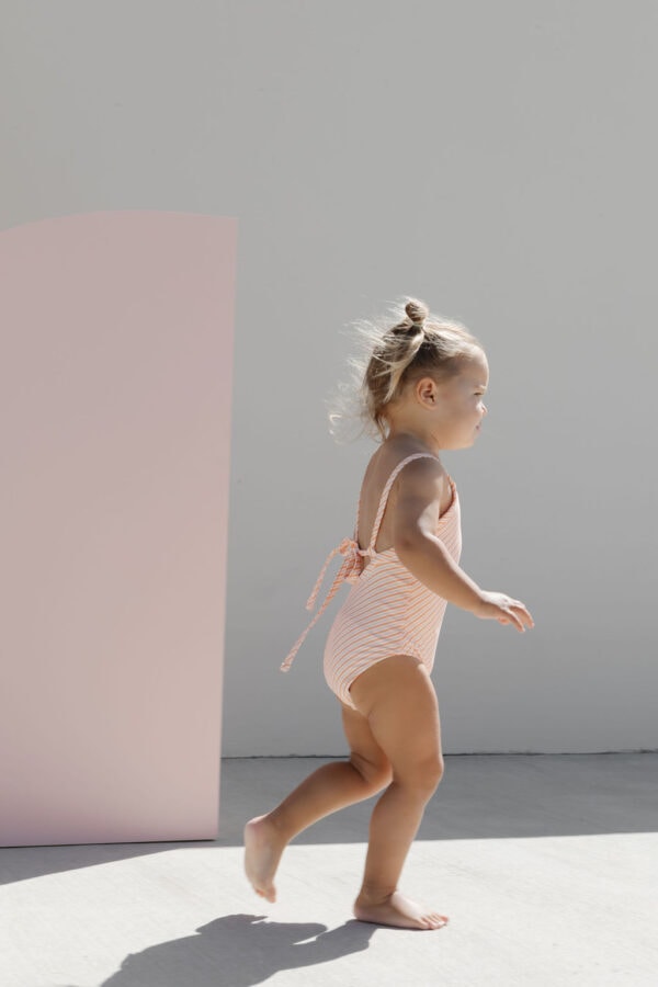 A toddler in a Mara One-Piece - Marigold Stripe swimsuit walks barefoot beside a pink board on a sunny day.