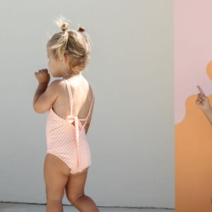 Toddler in a Mara One-Piece - Marigold Stripe swimsuit walking beside a wall with a graphic design.
