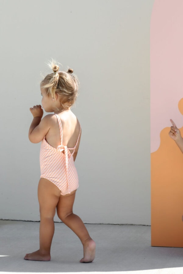 Toddler in a Mara One-Piece - Marigold Stripe swimsuit walking beside a wall with a graphic design.
