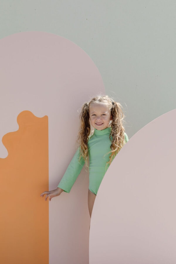 A young girl with curly hair peeking out from behind the June One-Piece - Fern on a wall.