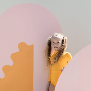 A young girl in a June One-Piece - Dandelion swimsuit smiling while peeking out from behind colorful abstract shapes.