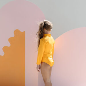 A child in a June One-Piece - Dandelion stands in front of a colorful backdrop with abstract shapes.