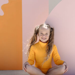 June One-Piece - Dandelion smiling child sitting cross-legged against a colorful backdrop.