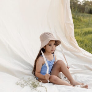 A little girl sitting on a blanket in a field wearing the WS - Golden Meadows Collection - Golden Sun Bucket Hat.