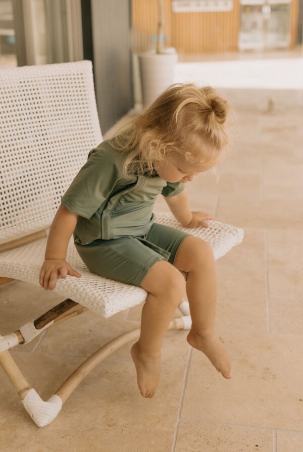 A little girl sitting on a Zimmi Onesie - Moss-covered white wicker chair.
