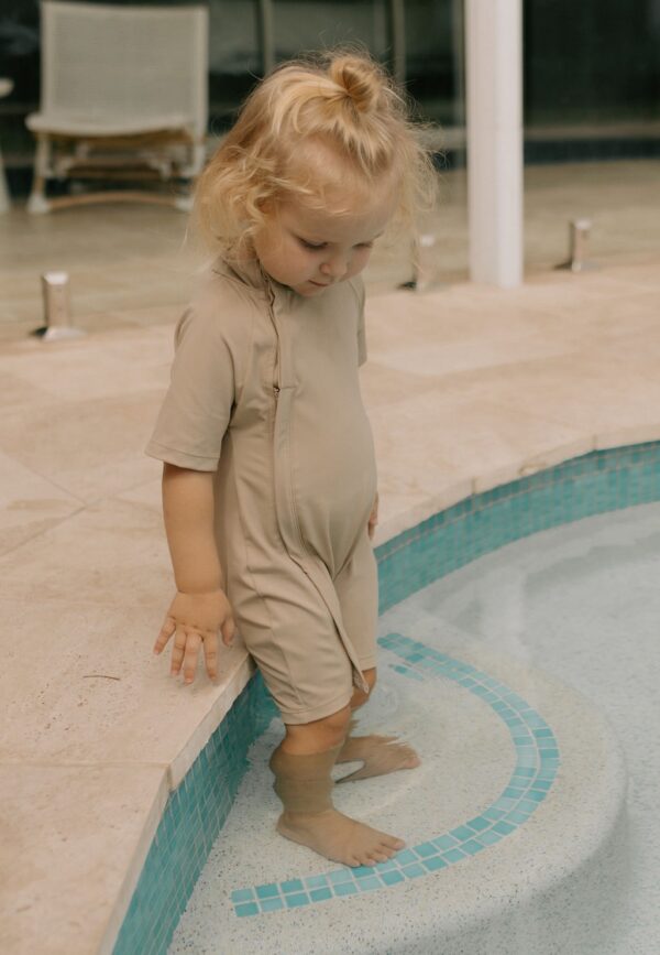 A little girl standing on the edge of a pool wearing an Essentials Range - Zimmi Onesie - Sand.