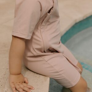 A child sitting on the edge of a pool wearing the Essentials Range - Zimmi Onesie - Rose swimsuit.