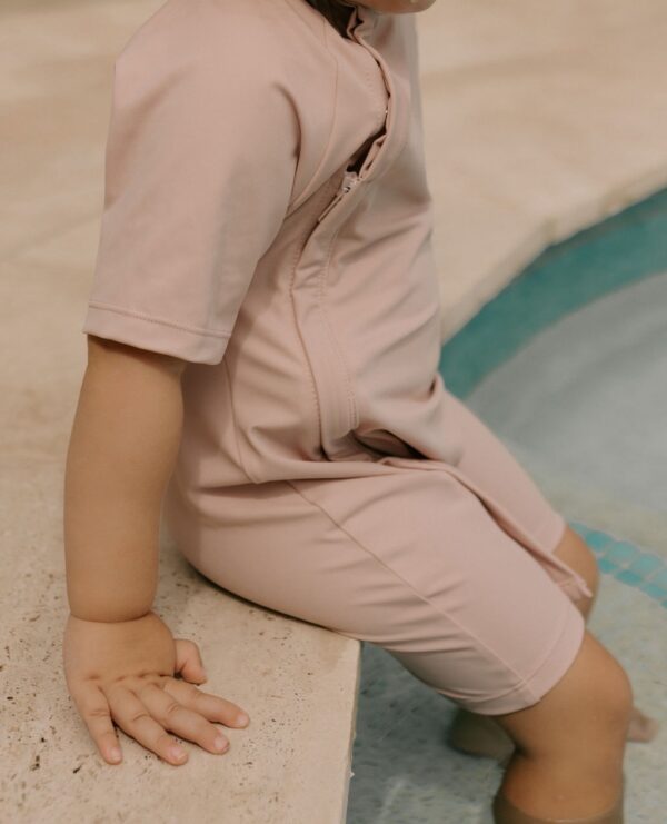 A child sitting on the edge of a pool wearing the Essentials Range - Zimmi Onesie - Rose swimsuit.