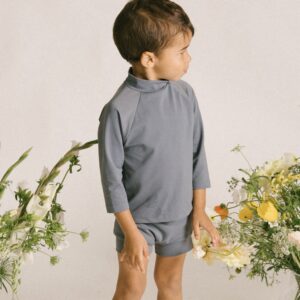 A young boy is standing in front of an Essentials Range - Nella Rash Shirt - Mineral Colour.