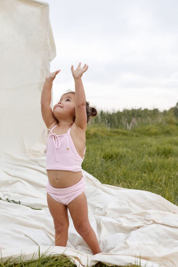 A young child in a Luna Bikini - Peach Blossom reaching up towards a billowing white cloth outdoors.