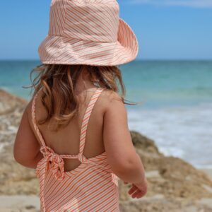 A little girl is standing on the beach wearing a Retro Wave By Ina - Mara One-Piece - Marigold Stripe.