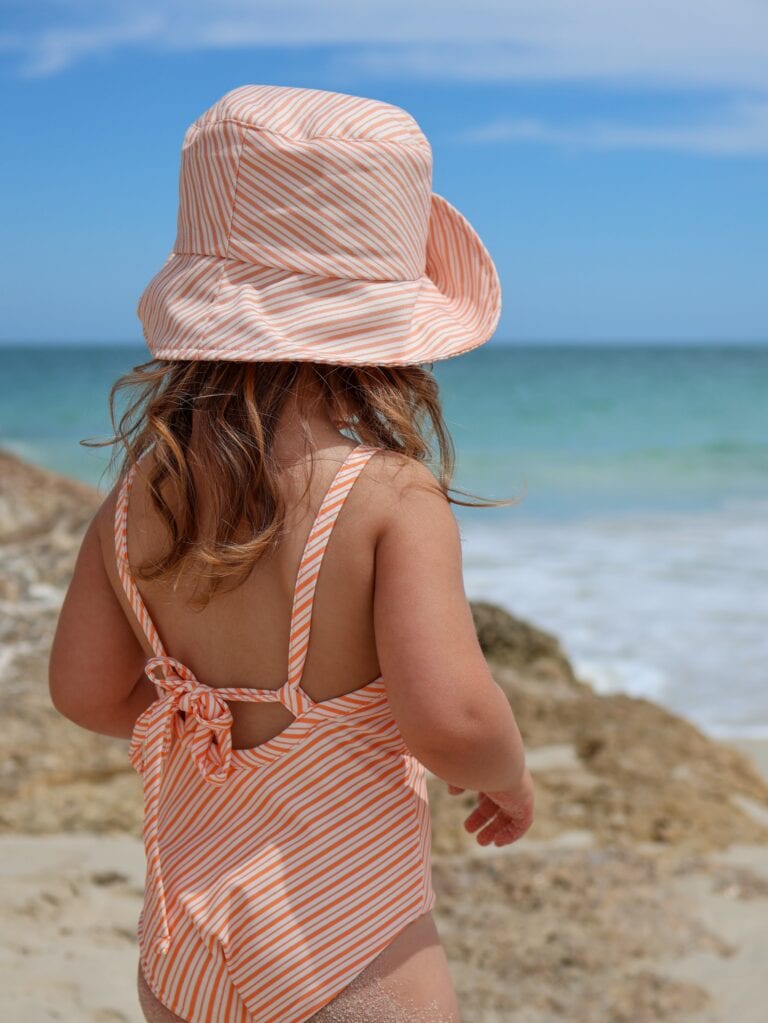A little girl is standing on the beach wearing a Retro Wave By Ina - Mara One-Piece - Marigold Stripe.