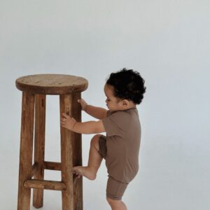 A baby is standing on a stool in front of a white background wearing the Essentials Range - Zimmi Onesie - Tort.